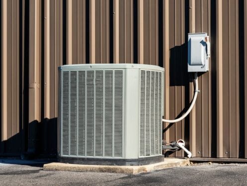 Outdoor residential air conditioning condenser unit installed on a concrete pad against a brown metal building wall.