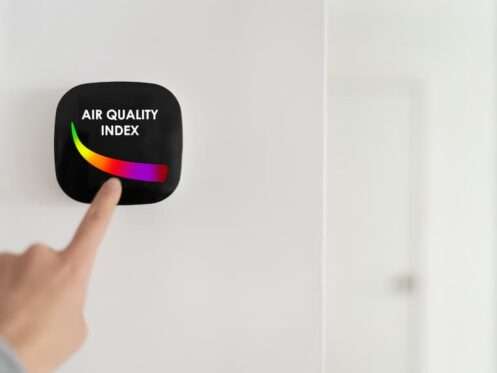 Hand touching a black digital screen mounted on a wall displaying an Air Quality Index color-coded gauge, highlighting the need for system maintenance to improve home indoor air quality.